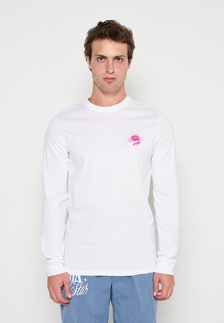 Junger Mann trägt ein weißes langärmliches Hemd mit kleinem pinkfarbenem Blumendesign auf der Brust und blaue Hose mit weißem Text.