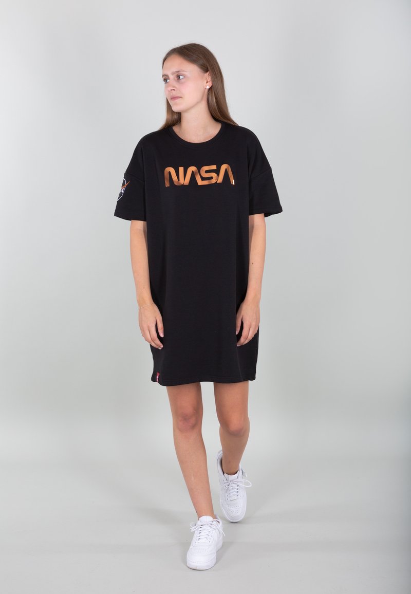Schwarzes, übergroßes T-Shirt-Kleid mit orangefarbenem NASA-Logo, kurzen Ärmeln und seitlichem Logo-Akzent. Getragen mit weißen Sneakern vor einem hellen Hintergrund.