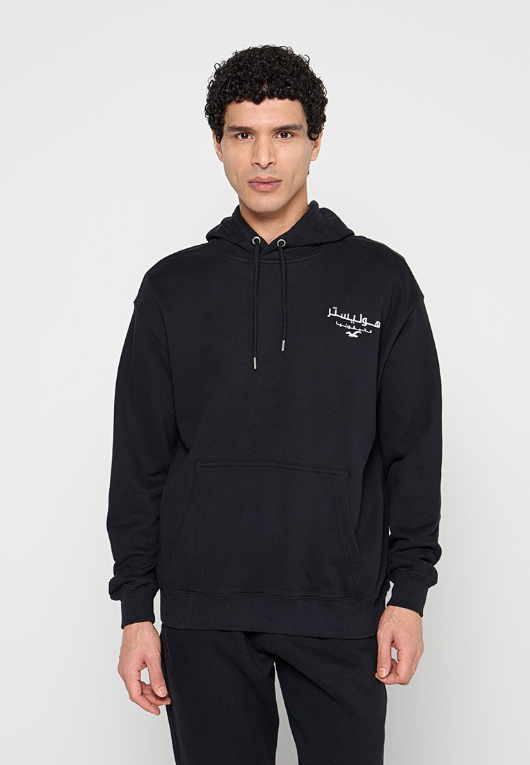Mann mit lockigem Haar, der einen schwarzen Hoodie mit weißer arabischer Schrift auf der Brust trägt und vor einem schlichten hellen Hintergrund steht.