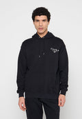 Mann mit lockigem Haar, der einen schwarzen Hoodie mit weißer arabischer Schrift auf der Brust trägt und vor einem schlichten hellen Hintergrund steht.