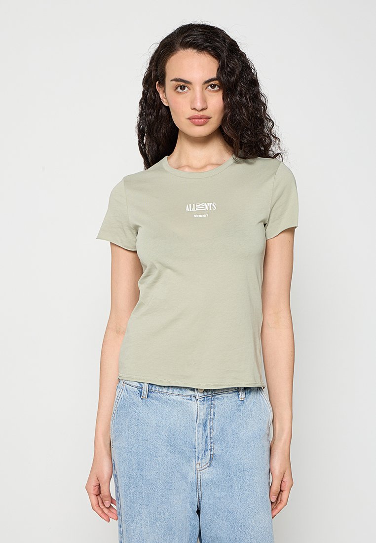 Frau mit lockigem dunklem Haar, die ein hellgrünes T-Shirt mit kurzen Ärmeln und weißem Text sowie hellblaue hoch taillierte Jeans trägt, steht vor einem weißen Hintergrund.