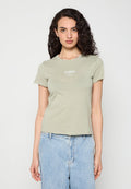 Frau mit lockigem dunklem Haar, die ein hellgrünes T-Shirt mit kurzen Ärmeln und weißem Text sowie hellblaue hoch taillierte Jeans trägt, steht vor einem weißen Hintergrund.