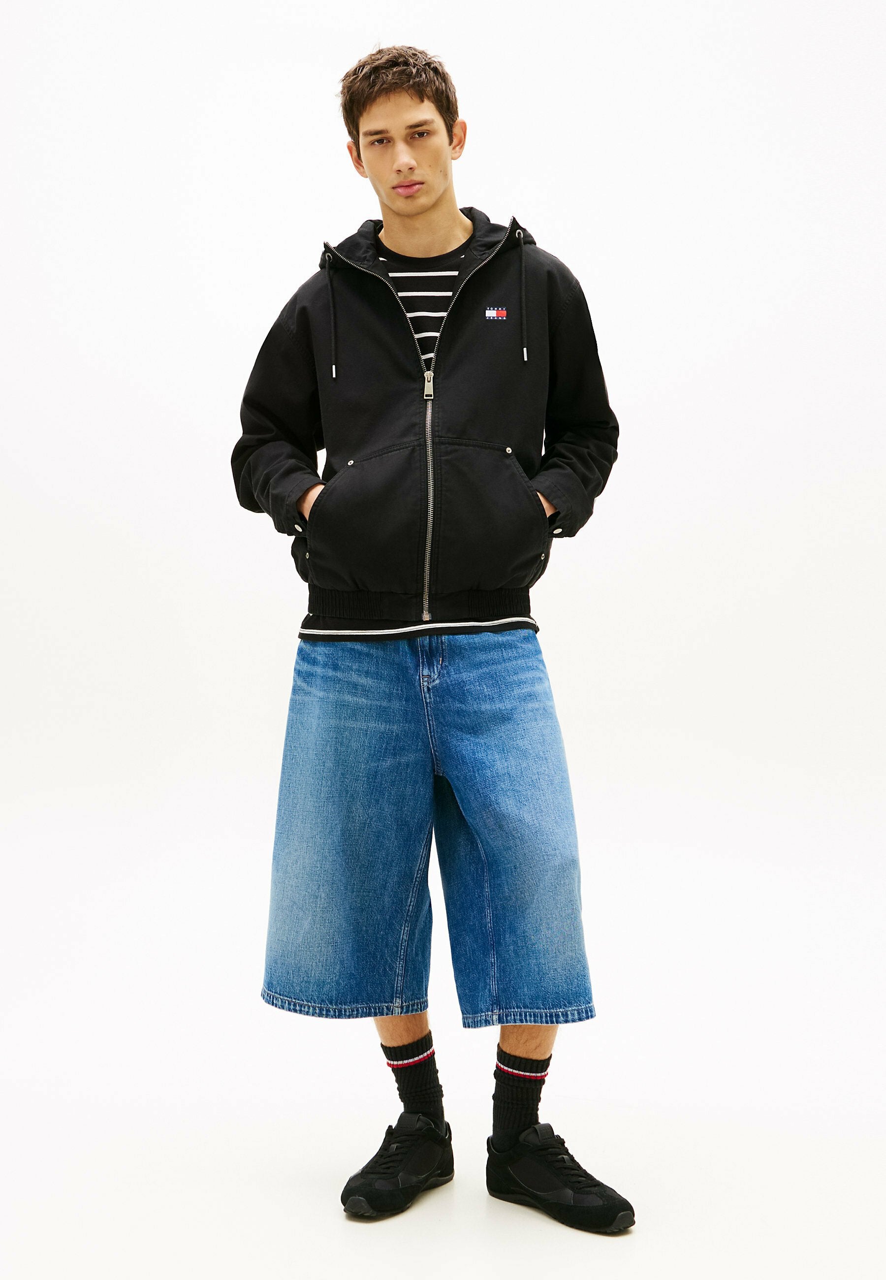 Junger Mann mit Händen in den Taschen, trägt einen schwarzen Hoodie mit Reißverschluss, ein gestreiftes Hemd, kniehohe Jeansshorts, schwarze Socken mit Streifen und schwarze Turnschuhe.