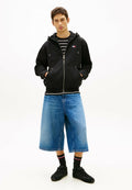 Junger Mann mit Händen in den Taschen, trägt einen schwarzen Hoodie mit Reißverschluss, ein gestreiftes Hemd, kniehohe Jeansshorts, schwarze Socken mit Streifen und schwarze Turnschuhe.