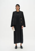 Schwarze taillierte Blazer mit goldenen Knöpfen über einem schwarzen Maxikleid. Das Model hält eine kleine weiße Tasche und trägt schwarze Schnürschuhe. Weißer Hintergrund.