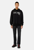 Schwarzes Hoodie mit einem ausgefransten weißen Logo, Kängurutasche, gerippte Bündchen, kombiniert mit dunklen Jeans und schwarzen Schuhen mit roten Akzenten.