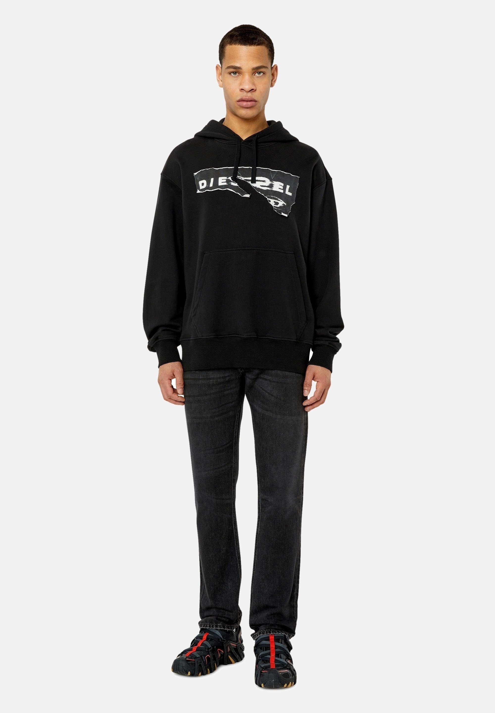 Schwarzes Hoodie mit einem ausgefransten weißen Logo, Kängurutasche, gerippte Bündchen, kombiniert mit dunklen Jeans und schwarzen Schuhen mit roten Akzenten.