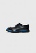 Schwarze Leder-Brogue-Schuhe mit blauer Sohlenstreifen und schwarzen Schnürsenkeln vor hellgrauem Hintergrund, Seitenansicht.