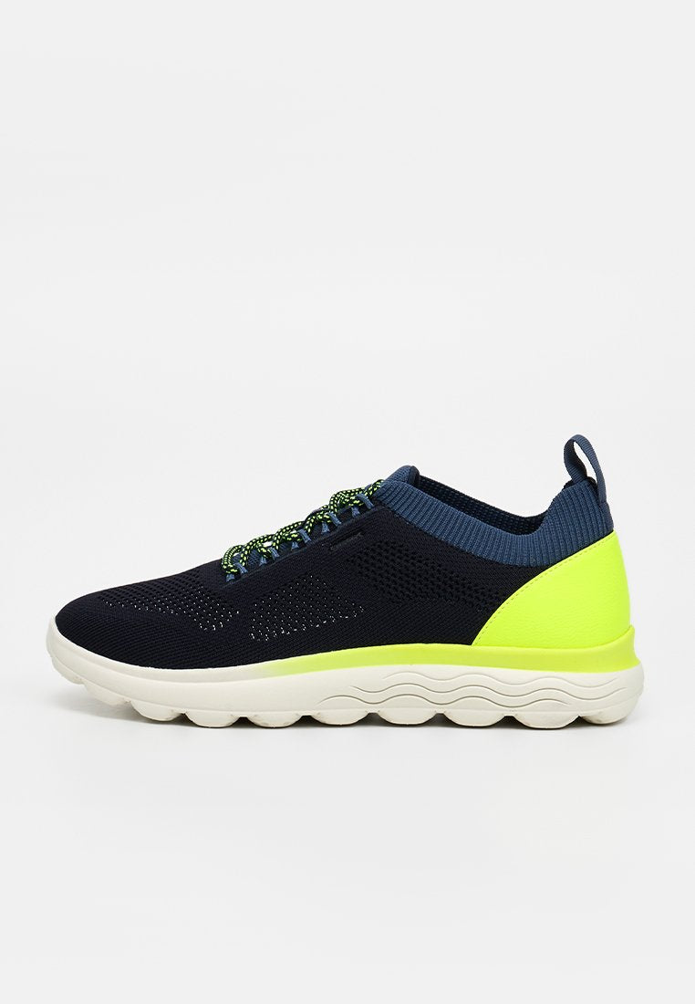 Navyblauer und neongelber Sneaker mit gestricktem Obermaterial, gepolsterter weißer Sohle und gemusterten Schnürsenkeln, von der Seite auf einfarbigem Hintergrund dargestellt.