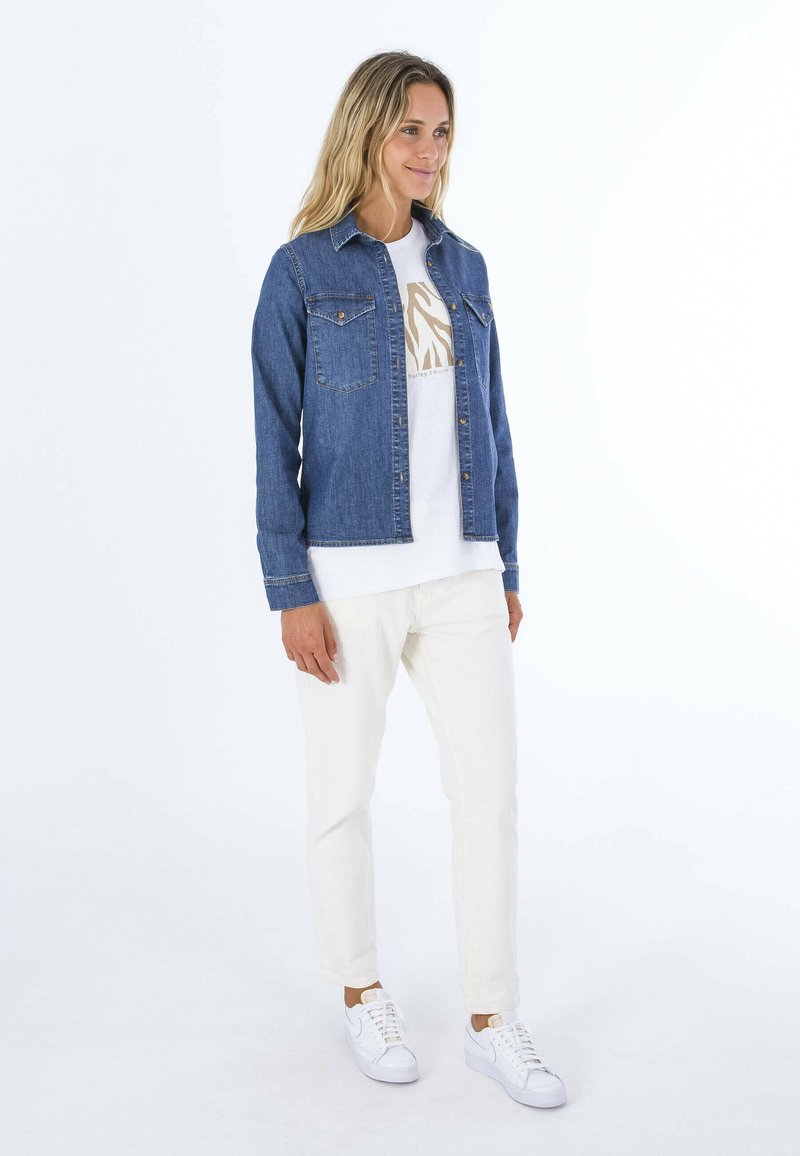 Jeansjacke mit zwei vorderen Taschen, getragen über einem weißen T-Shirt mit einem leichten Grafikdesign. Kombiniert mit weißen Hosen und Sneakers.