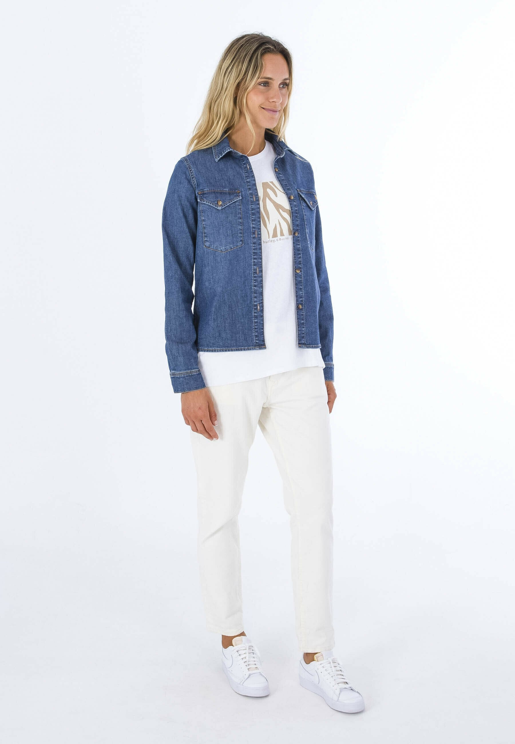 Jeansjacke mit zwei vorderen Taschen, getragen über einem weißen T-Shirt mit einem leichten Grafikdesign. Kombiniert mit weißen Hosen und Sneakers.