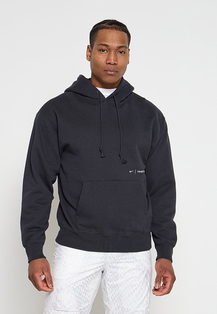 Junger Mann, der einen schwarzen Nike-Hoodie mit dem Schriftzug "Together" und weiße gemusterte Hose trägt, steht vor einem schlichten hellen Hintergrund.