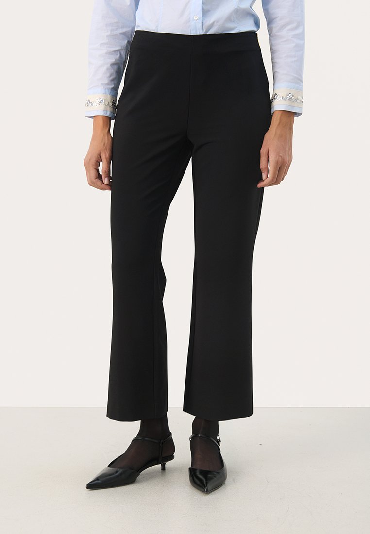 Schwarze, taillierte Hose aus glattem Stoff, mit einer figurschmeichelnden Taille und ausgestellten Beinen. Kombiniert mit spitzen schwarzen Schuhen.
