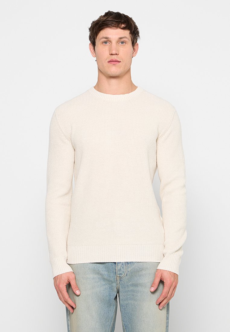 Cremefarbener, gerippter Strickpullover mit rundem Ausschnitt, langen Ärmeln und enganliegenden Bündchen, kombiniert mit hellblauen Denim-Jeans.