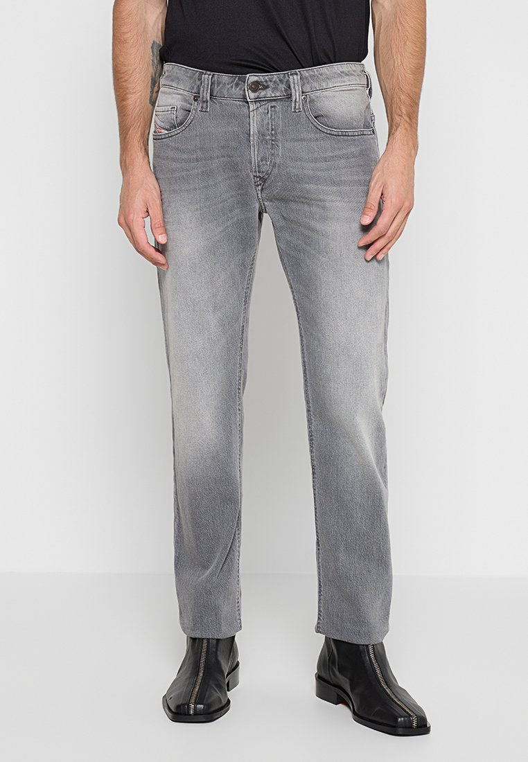 Graue Jeans aus Denim mit einem verblassten Finish, geradem Bein, Fünf-Taschen-Design und Metallbeschlägen. Getragen mit einem schwarzen enganliegenden Hemd und Stiefeln.