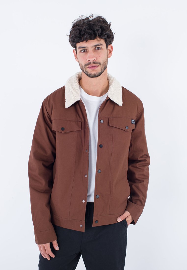 Braune Jacke mit einem cremefarbenen Lammfellkragen, Druckknöpfen, zwei Brusttaschen, glattem Stoff und einer lockeren Passform vor einem weißen Hintergrund.