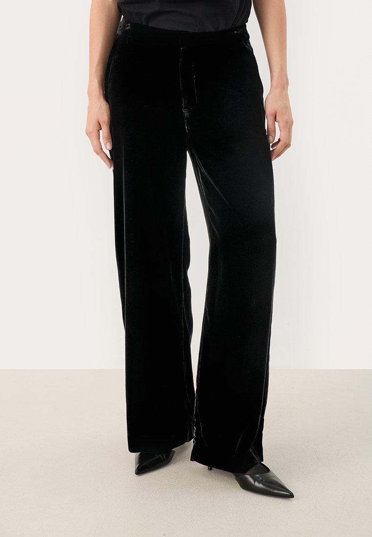 Schwarze Samt-Hose mit weitem Bein und glatter Textur, ausgestattet mit einer taillierten Taille und dezenten Vorderfalten. Tragen mit spitzen Absatzschuhen.
