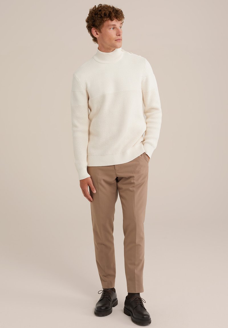 Cremfarbene, gerippte Strickjacke mit hochgeschlossenem Kragen, kombiniert mit hellbraunen, maßgeschneiderten Hosen und schwarzen Schnürschuhen. Lässiges Outfit.