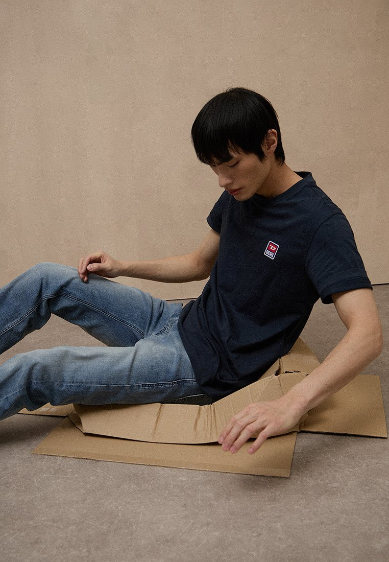 Marineblaues Baumwoll-T-Shirt mit einem kleinen Logo auf der Brust, kombiniert mit hellblauen Denim-Jeans, sitzend auf einem Kartonstück vor neutralem Hintergrund.