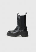 Schwarze Knöchelstiefel mit elastischen Seitenpaneelen und Zuglaschen, ausgestattet mit einer klobigen Sohle mit der Aufschrift "TOMMY." Einfaches, elegantes Design vor einem weißen Hintergrund.