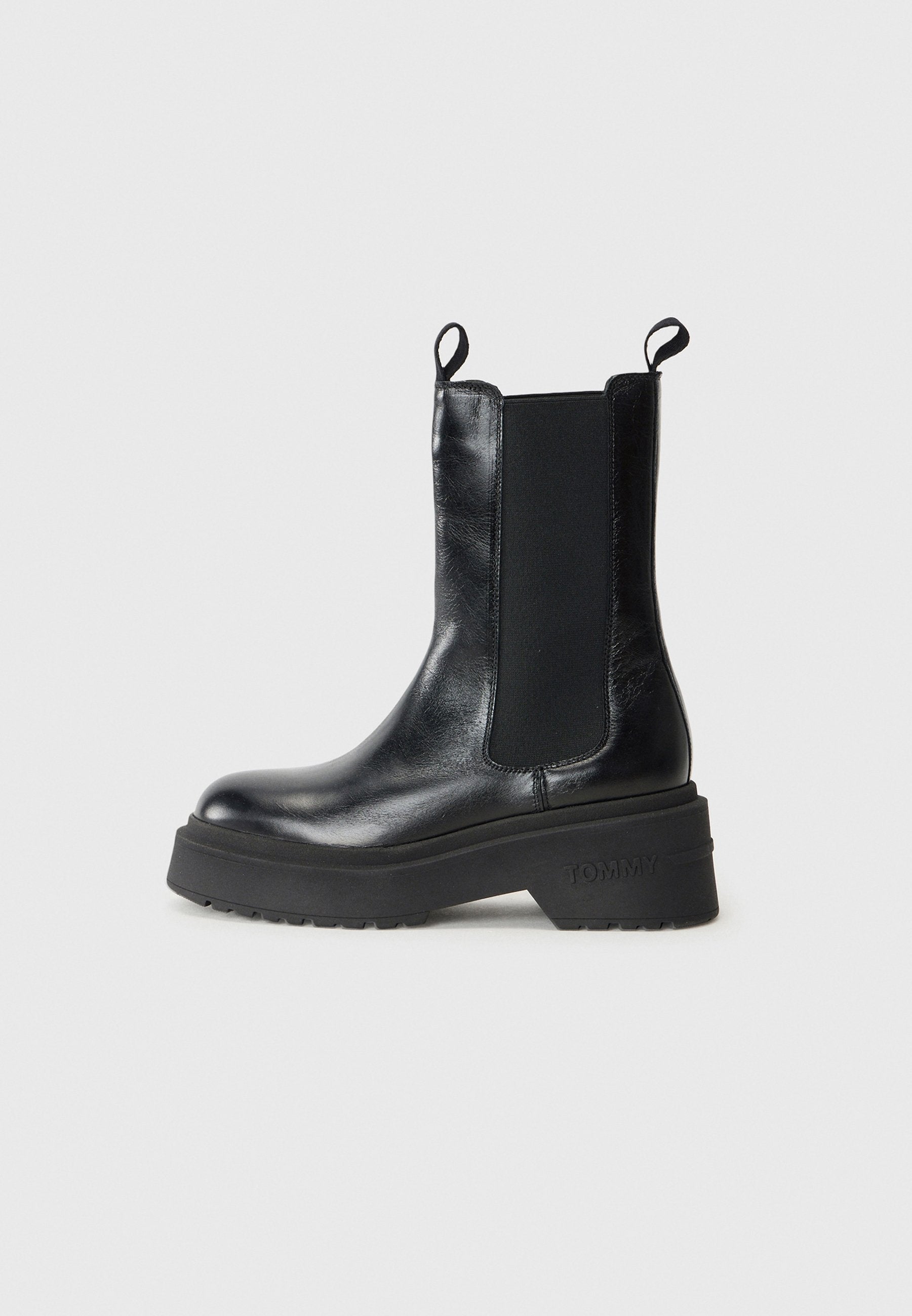 Schwarze Knöchelstiefel mit elastischen Seitenpaneelen und Zuglaschen, ausgestattet mit einer klobigen Sohle mit der Aufschrift "TOMMY." Einfaches, elegantes Design vor einem weißen Hintergrund.