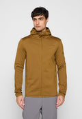 Mann trägt eine braune Kapuzenjacke mit Reißverschluss und graue Hose, steht vor einem einfarbigen hellgrauen Hintergrund.