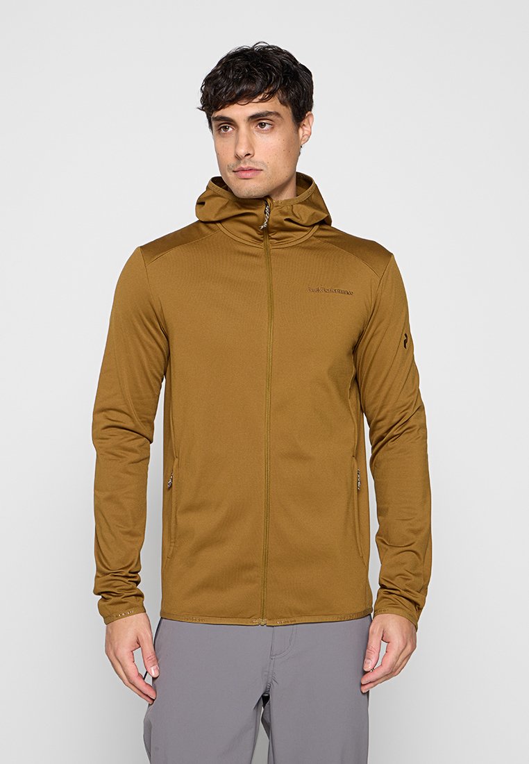 Mann trägt eine braune Kapuzenjacke mit Reißverschluss und graue Hose, steht vor einem einfarbigen hellgrauen Hintergrund.