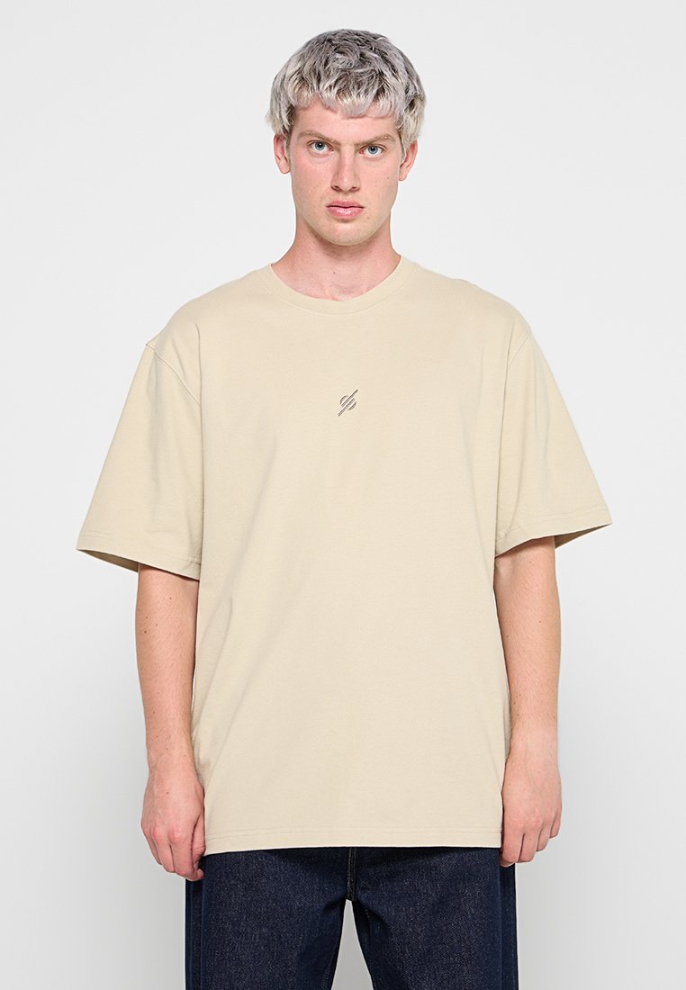 Junger Mann mit kurzen blonden Haaren, der ein beiges übergroßes T-Shirt mit einem kleinen Logo auf der Brustmitte und dunkelblauen Jeans trägt, aufrecht stehend.