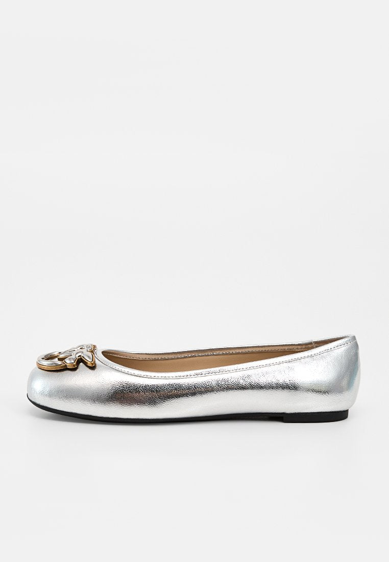 Silberfarbener metallischer Ballerina-Schuh mit schwarzer Sohle und goldenem dekorativem Emblem an der abgerundeten Spitze, dargestellt vor weißem Hintergrund.