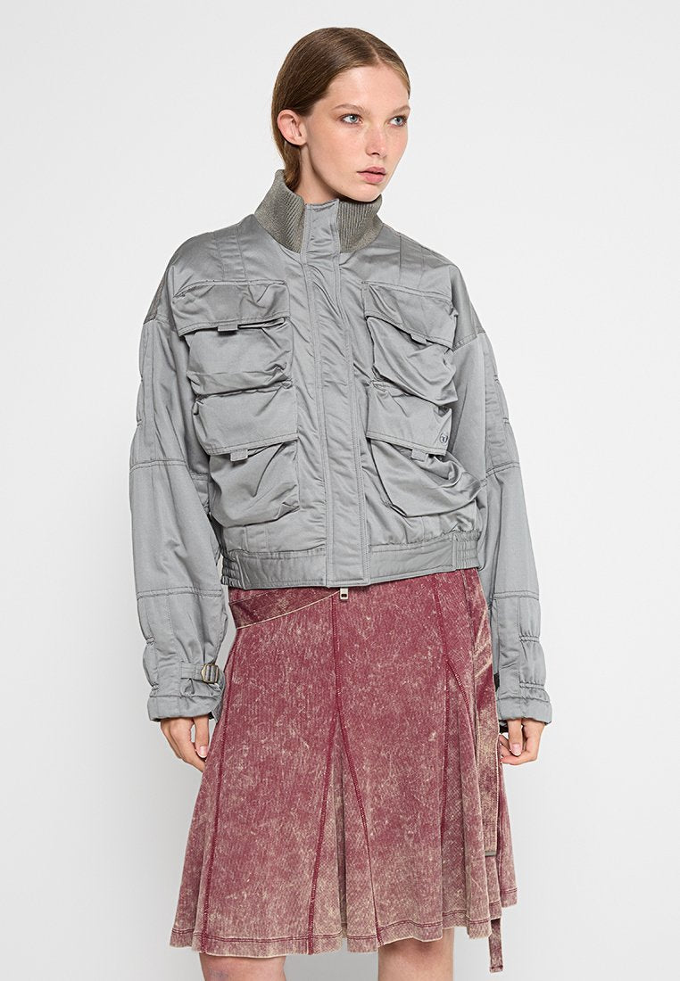 Graue Bomberjacke mit mehreren Fronttaschen, geripptem Kragen und elastischen Bündchen, kombiniert mit einem verblassten rosa Rock mit ausgestelltem Design.