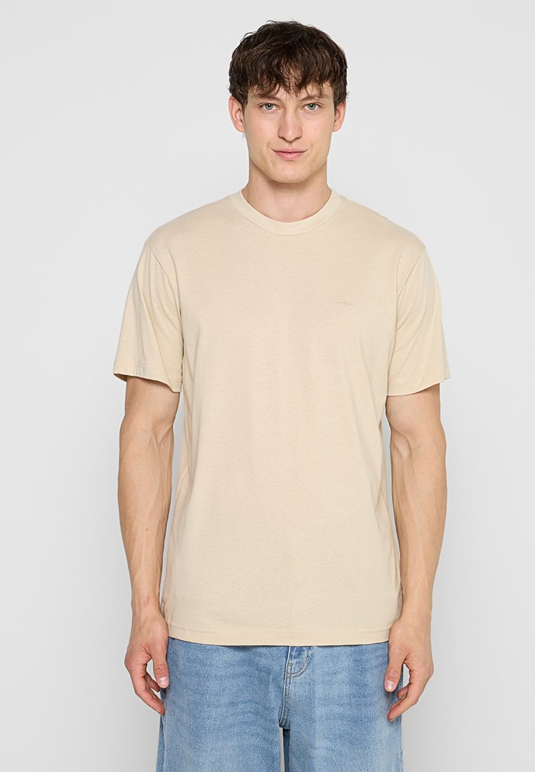 Junger Mann mit kurzen braunen Haaren, der ein beiges T-Shirt mit kurzen Ärmeln und hellblaue Jeans trägt, steht vor einem einfarbigen hellgrauen Hintergrund.