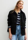 Schwarze Jacke mit Reißverschluss, zwei frontalen Taschen und einem Kragen. Getragen über einem blau-weißen gestreiften Hemd und hochtaillierten blauen Jeans.