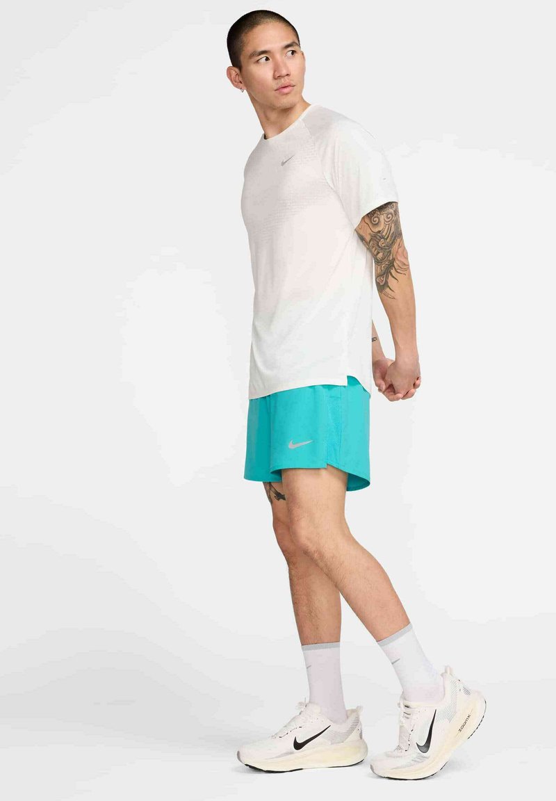 Helles graues Sport-T-Shirt mit kurzen Ärmeln, kombiniert mit türkisfarbenen Shorts und weißen Laufschuhen. Enthält Nike-Logos auf der Bekleidung.