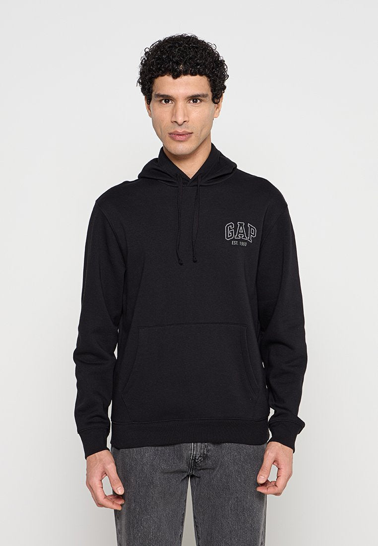 Mann mit lockigem Haar, der einen schwarzen GAP-Hoodie mit Fronttasche und graue Jeans trägt, vor einem einfarbigen hellen Hintergrund stehend.