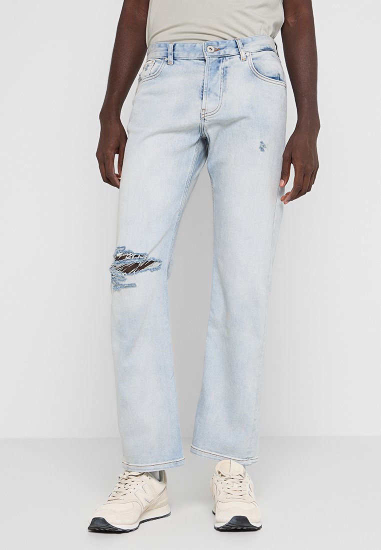 Helle blaue Jeans mit lockerem Schnitt, mit Rissen am linken Knie und einem klassischen Fünf-Taschen-Design. Getragen mit beigen Sneakern.