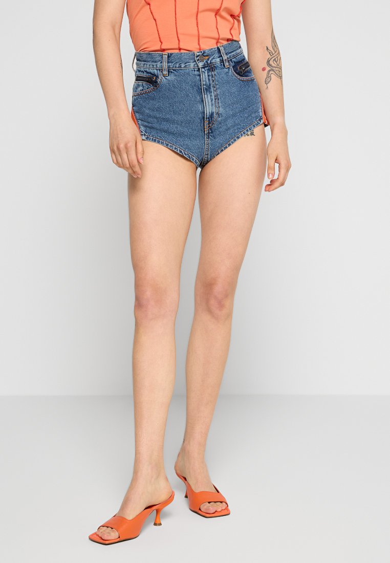 Blaue High-Waist-Jeansshorts mit fransigem Saum, kombiniert mit einem orangefarbenen Oberteil und orangefarbenen Absatzschuhen. Glatte Denim-Oberfläche sichtbar.