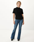 Schwarzer kurzärmliger Pullover mit hohem Kragen, kombiniert mit weit geschnittenen blauen Jeans und schwarzen Stiefeln mit Absatz, vor einem minimalistischen weißen Hintergrund.