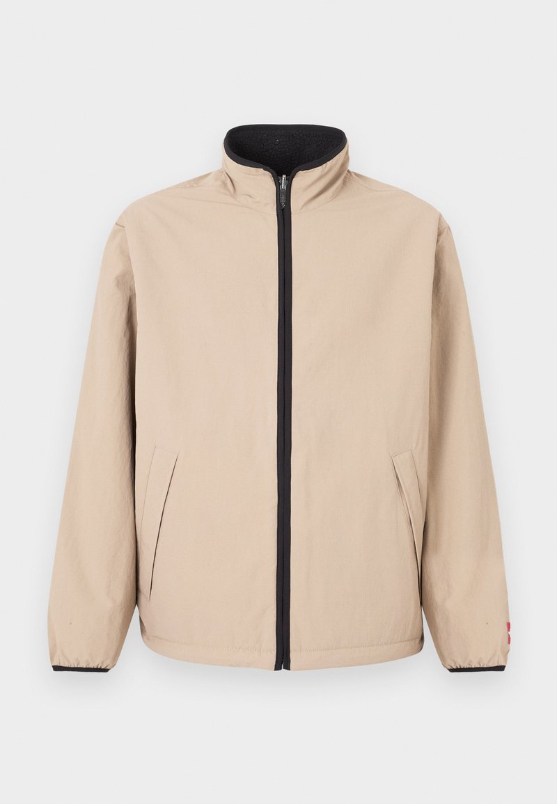 Beige Jacke mit hohem Kragen, Reißverschluss vorne und schwarzen Akzenten. Verfügt über zwei Seitentaschen und elastische Bündchen. Glatte Textur.