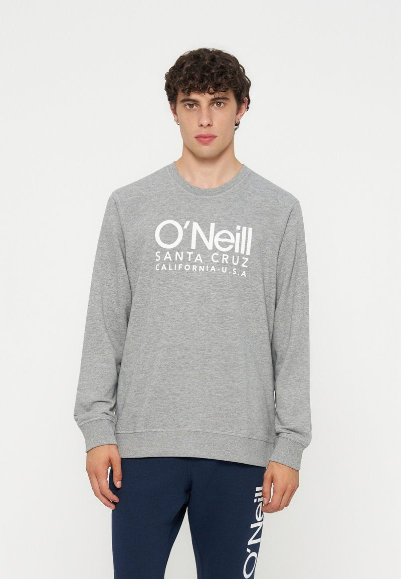 Junger Mann mit lockigem Haar, der einen grauen O'Neill Langarm-Sweatshirt und marineblaue O'Neill Hose trägt, steht vor einem schlichten Hintergrund.