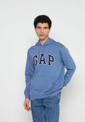 Mann mit lockigem Haar, der einen blauen GAP-Hoodie und blaue Jeans trägt, steht mit einer Hand hinter dem Rücken vor einem schlichten weißen Hintergrund.