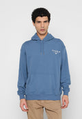Mann trägt blauen Kapuzenpullover mit arabischem Text und Logo auf der Brust, steht vor einfarbigem hellgrauem Hintergrund.