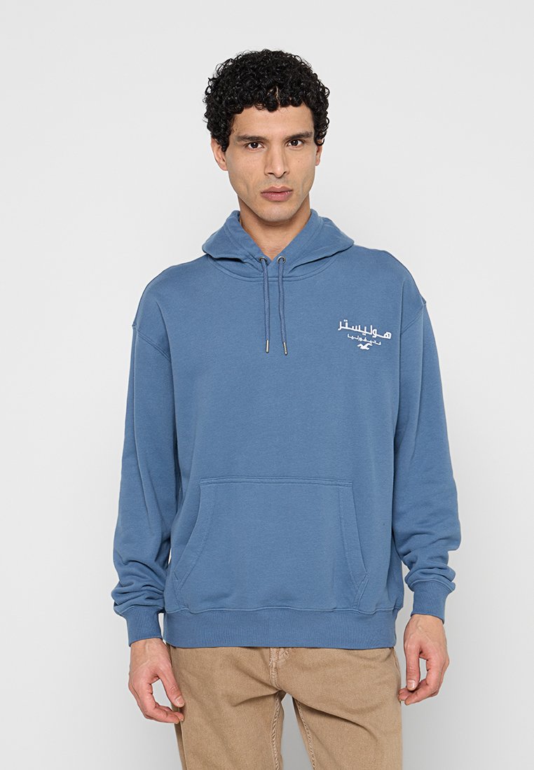 Mann trägt blauen Kapuzenpullover mit arabischem Text und Logo auf der Brust, steht vor einfarbigem hellgrauem Hintergrund.