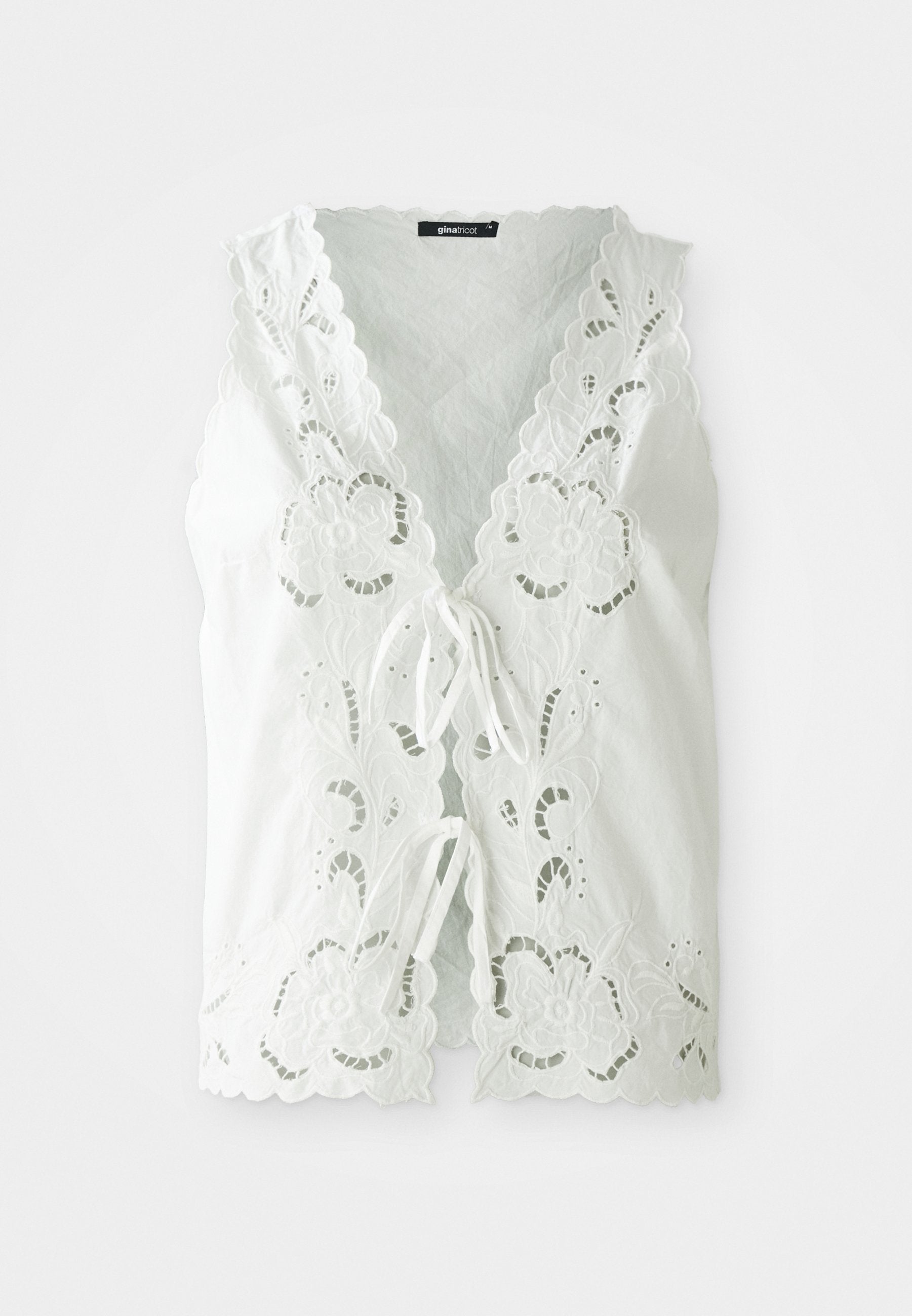 Weiße ärmellose Bluse mit floralen Spitzenausschnitten, gewellten Kanten und zwei vorderen Bindeschließen, vor einem schlichten Hintergrund.