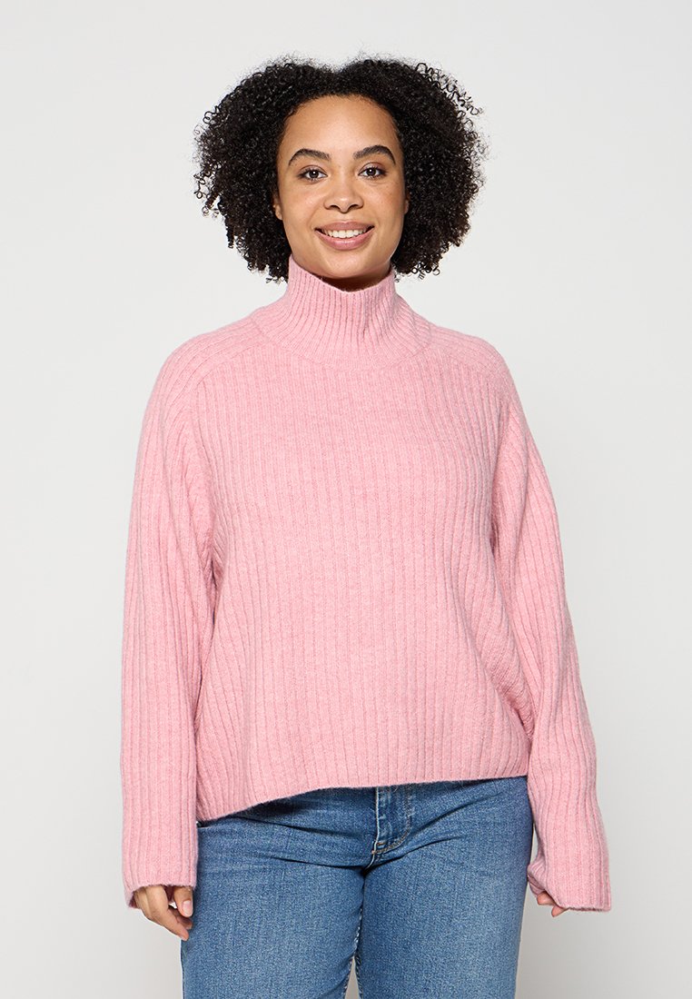 Lächelnde Frau mit lockigem Haar, die einen gerippten pinken Rollkragenpullover und blaue Jeans trägt, vor einem einfarbigen hellen Hintergrund stehend.