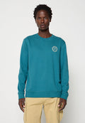 Person trägt ein dunkelgrünes Sweatshirt mit langen Ärmeln und kleinem hellblauem Logo auf der Brust sowie beige Cargohosen und steht vor einem einfarbigen Hintergrund.