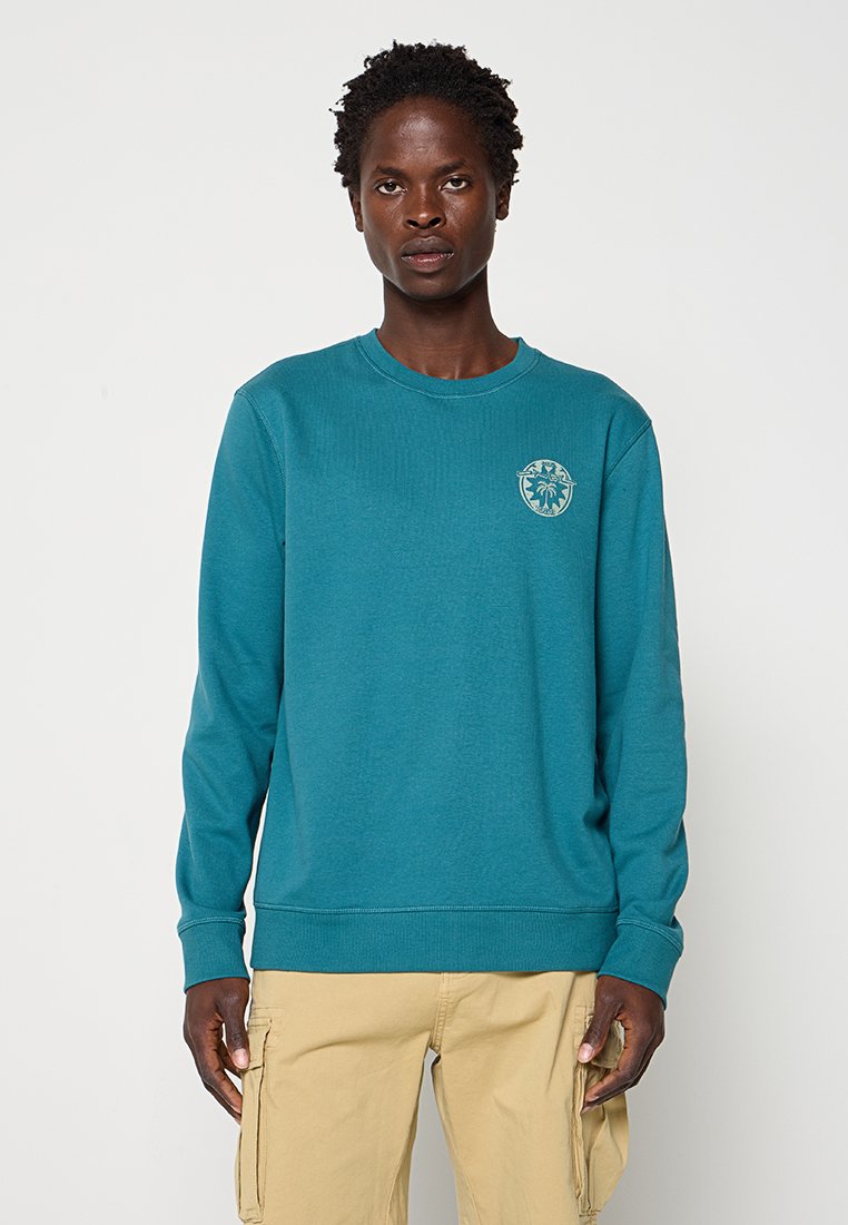 Person trägt ein dunkelgrünes Sweatshirt mit langen Ärmeln und kleinem hellblauem Logo auf der Brust sowie beige Cargohosen und steht vor einem einfarbigen Hintergrund.