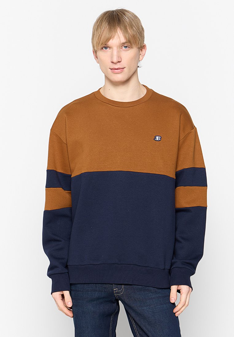Junger Mann mit kurzen blonden Haaren, der einen sweatshirt in braun und marineblau mit Farbschnitt und dunkelblauen Jeans trägt, vor einem weißen Hintergrund steht.