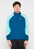 Junger Mann, der eine blau-mintgrüne Nike Sportjacke mit Reißverschluss und schwarze Netz-Shorts trägt, steht frontal vor einem schlichten Hintergrund.