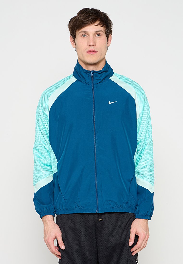 Junger Mann, der eine blau-mintgrüne Nike Sportjacke mit Reißverschluss und schwarze Netz-Shorts trägt, steht frontal vor einem schlichten Hintergrund.