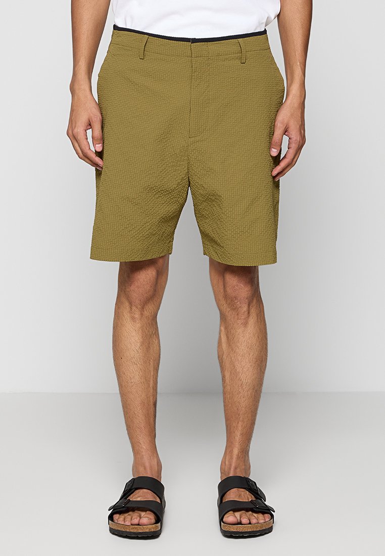 Olivgrüne Shorts mit einer strukturierten Oberfläche, knielang, mit einem schwarzen Gummibund und getragen mit schwarzen Sandalen.